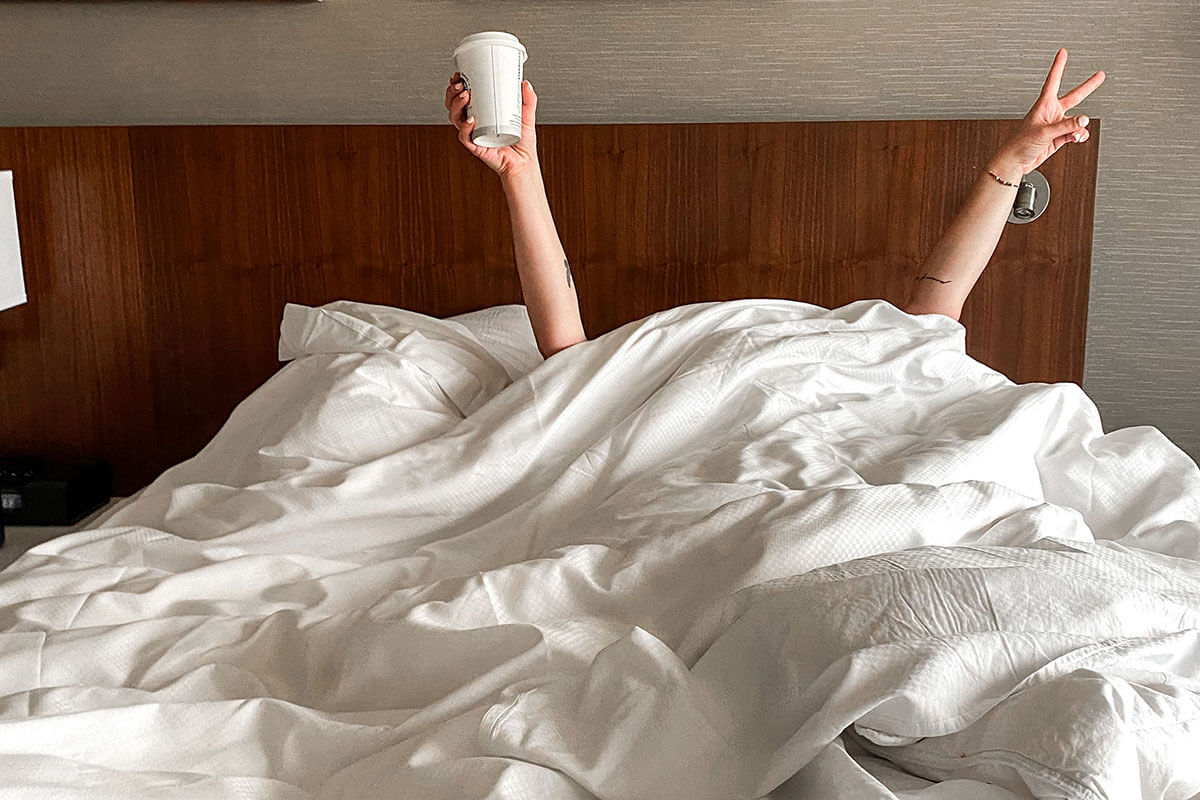 A girl under the bed covers celebrating with a cup of takeout coffee, decompressing in the Regina January City Guide