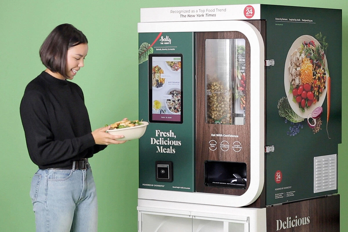 A girl standing beside a Chowbotics’ Sally 2.0 machine with her freshly made salad