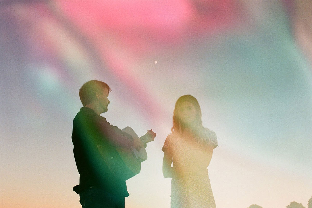 One of 5 up-and-coming Canadian musicians, Ocie Elliott is a female-male duo, here standing against a colourful sky with guitar ready