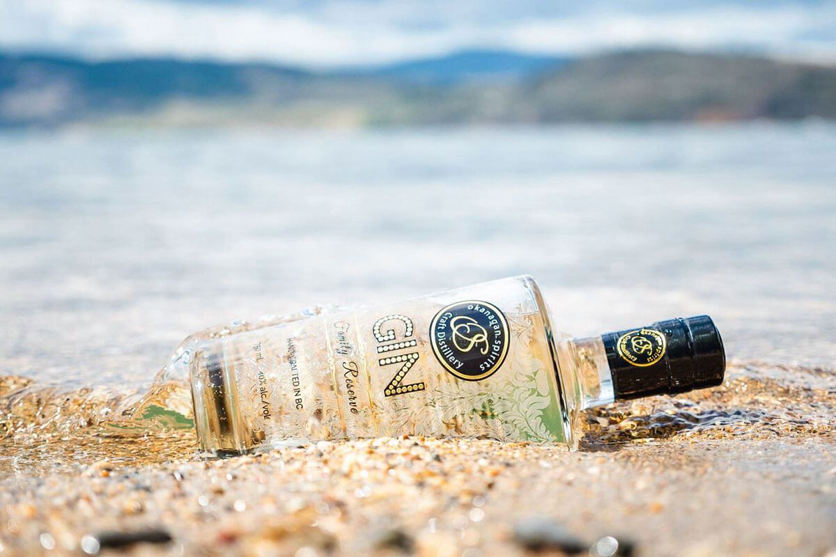 A bottle of Okanagan Spirits Craft Distillery Family Reserve Gin laying in the sand on the edge of a lake, part of  food & wine in Kelowna