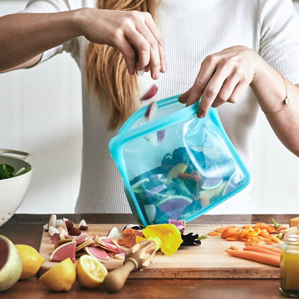 A woman holding one of the most popular plastic-free storage products - a blue Stasher bag from EarthHero, and filling it with fresh salad ingredients