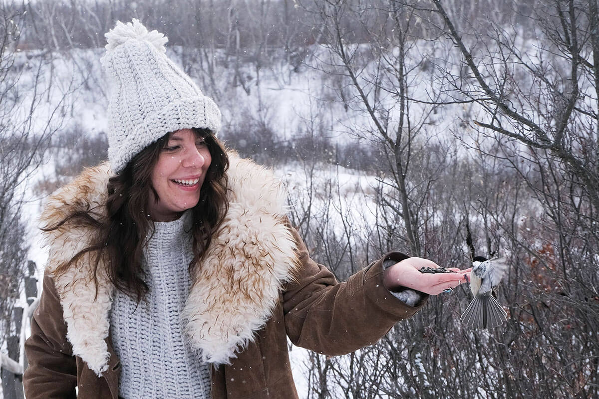 A woman outside in winter with a chickadee brid eating from her hand  is part of the Saskatoon February City Guide