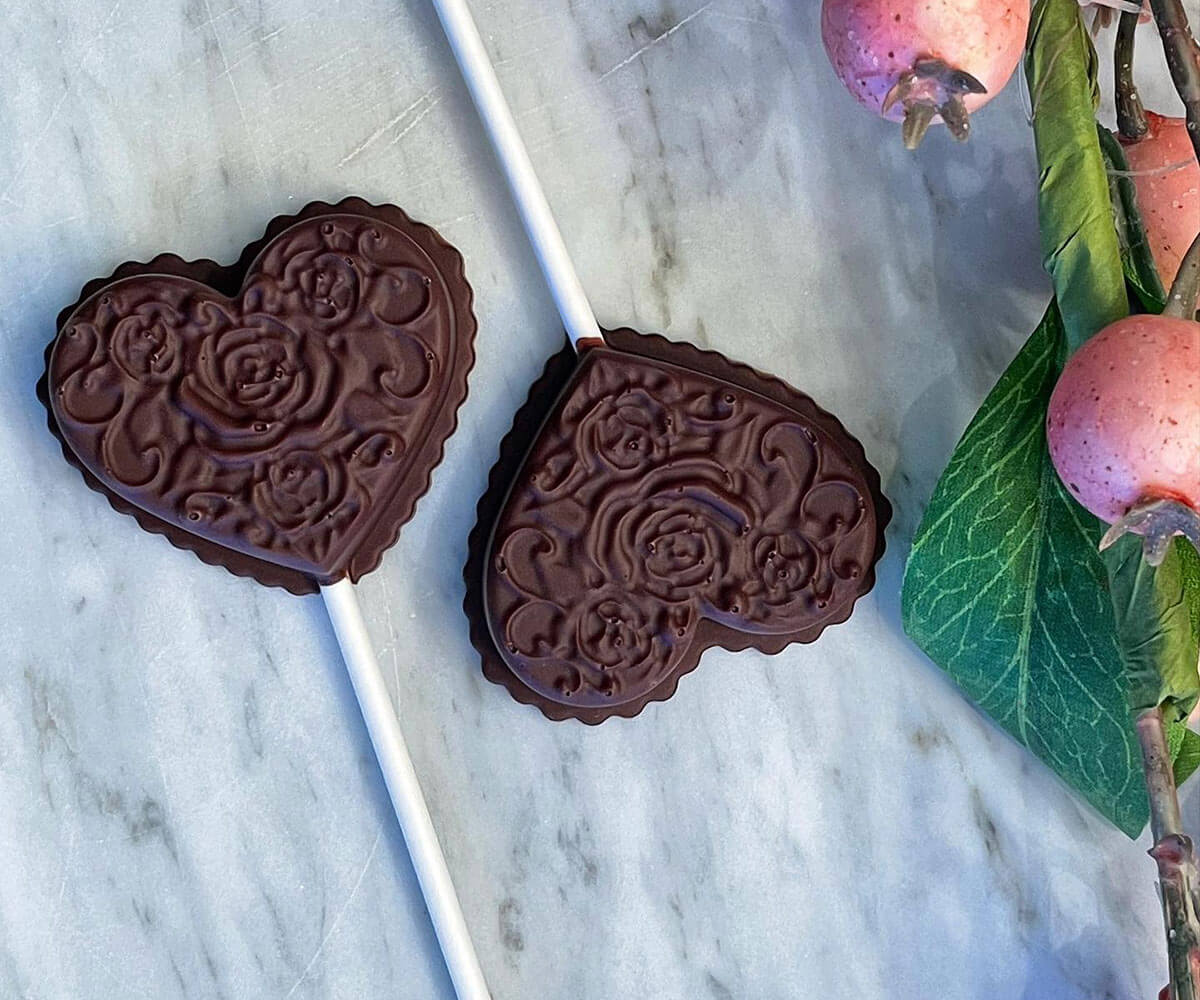 Two heart-shaped chocolates on white sticks beside link berries on a granite counter