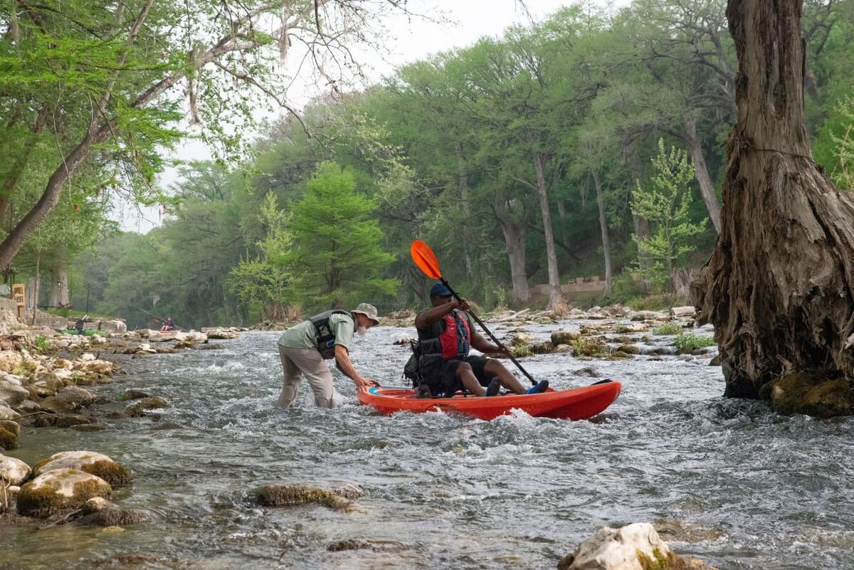 man in kayak on river surrounded by forest being helped by another man, part of Heroes on the Water