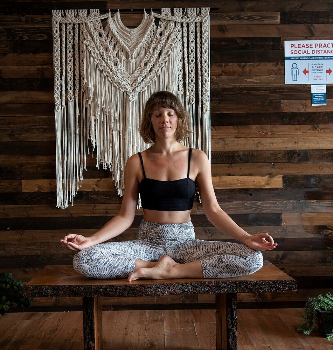 A woman meditating on a wooden bench with a macrame hanging behind her