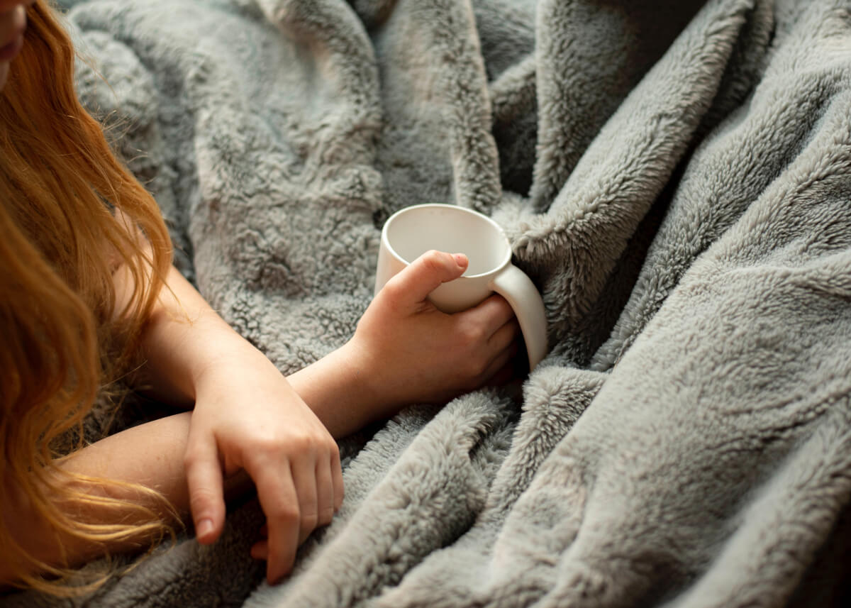 person with long red hair holding a white mug under a weighted blanket, one of many natural anxiety remedies