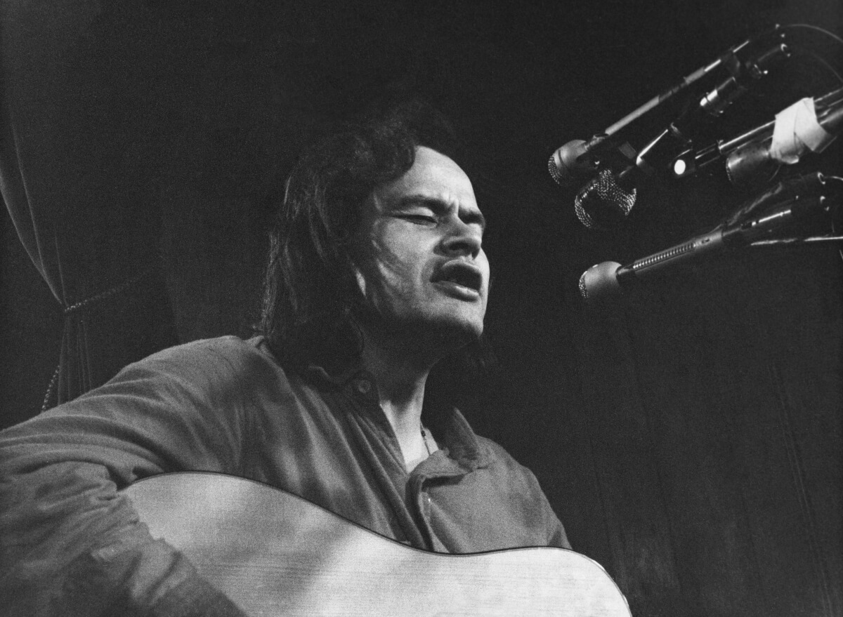 Black and white photo of Willie Dunn singing and playing guitar, 1976