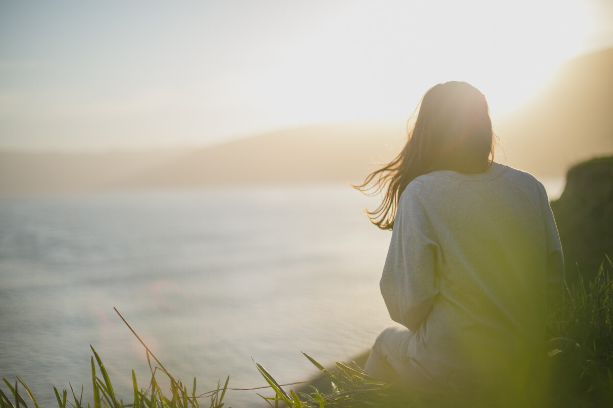 A person looks serene sitting in the grass looking out over the water. Ten Percent Happier is an app for mental wellness. 