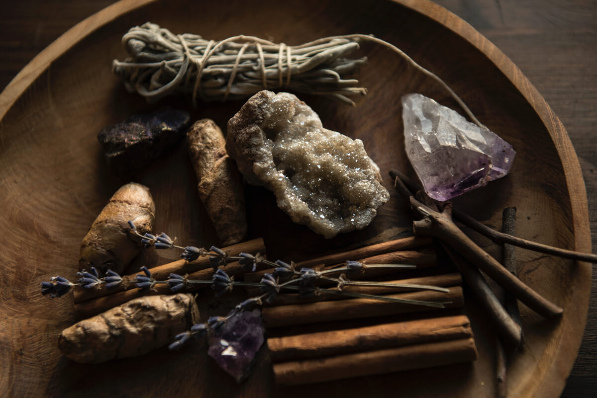 Ingredients for an urban witch spell. Cinnamon sticks, amethyst, lavender, ginger, and other items are pictured in a wooden bowl. 