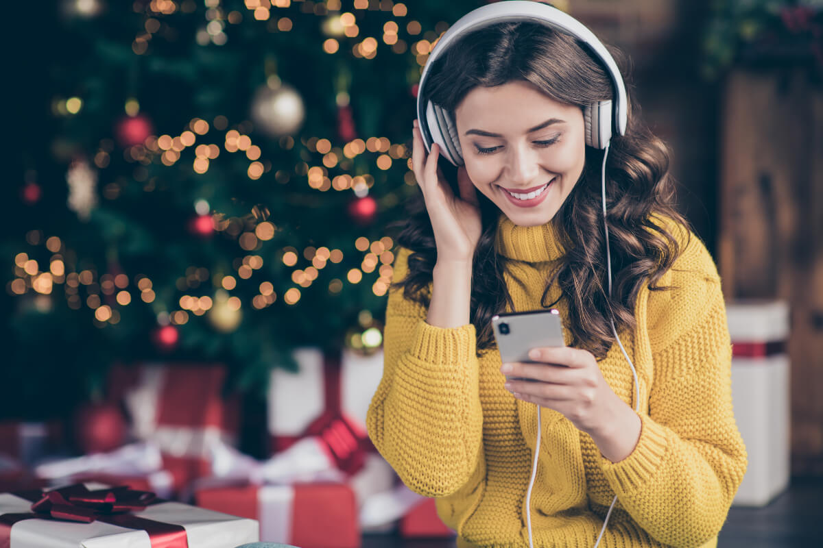 Woman smiles while listening to her holiday playlist on her headphones (with a Christmas tree in the background).