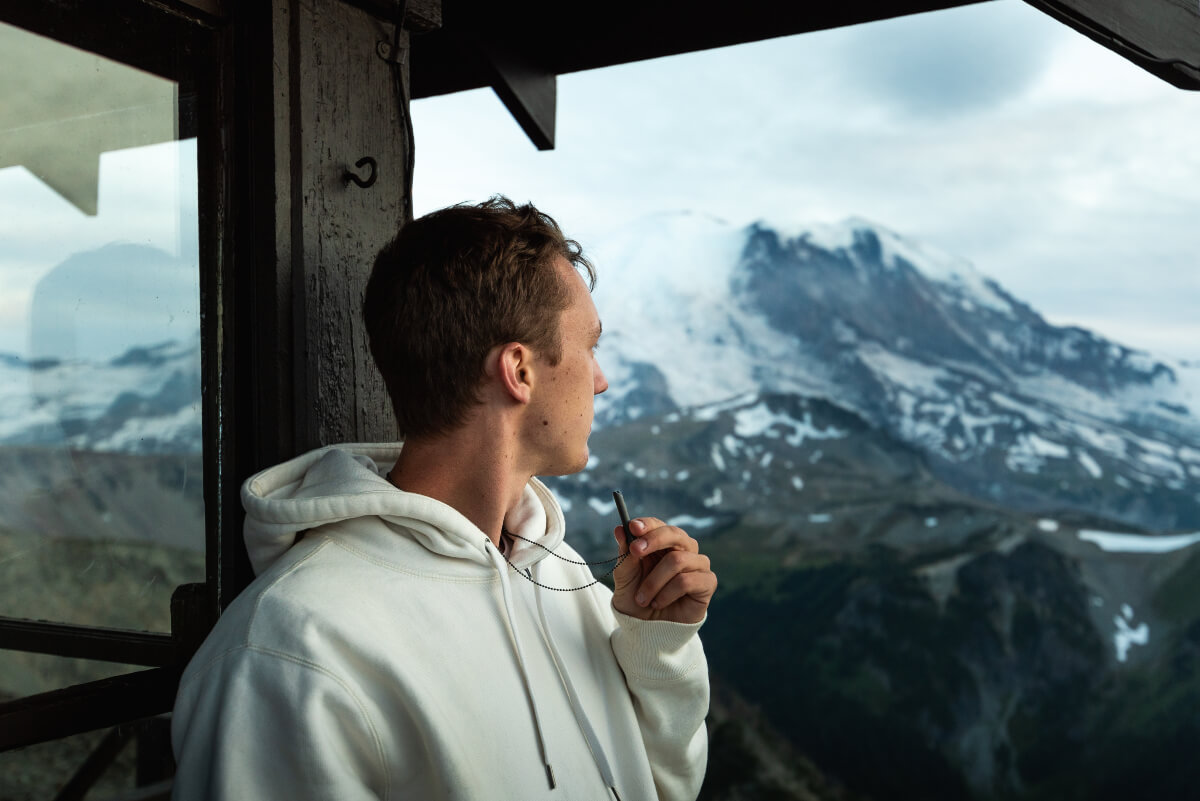 A man holds his shift necklace while looking out at a view of a snow-capped mountain. 