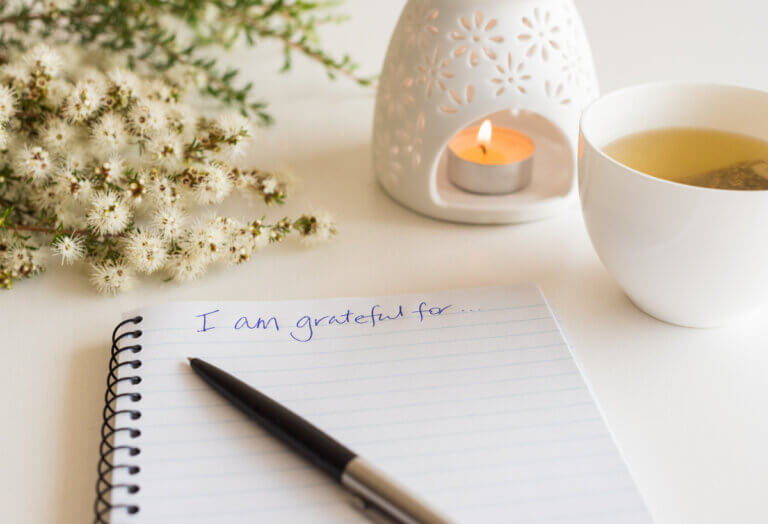 "I am grateful for:" is written at the top of a notepad surrounded by a white candle and white flowers.