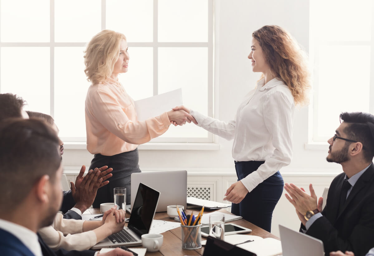 Two colleagues shaking hands and smiling at a business meeting while colleagues around them clap. Setting good goals includes being specific. 