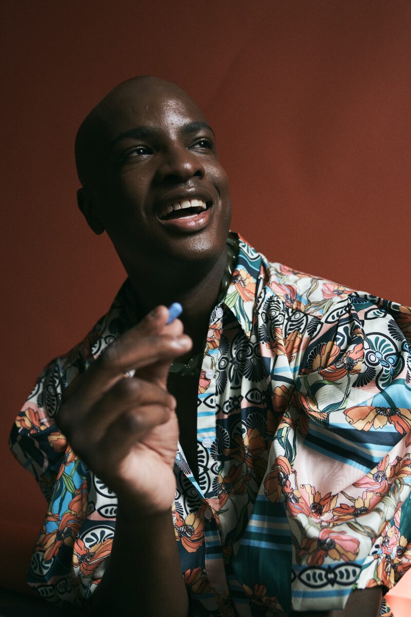 Person smiling holding a blue pill while wearing a funky-patterned shirt. 