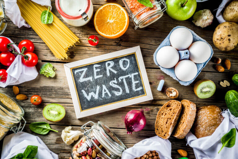 "Zero Waste" written on a mini chalk board surrounded by eggs, bread, fruit, and other staples
