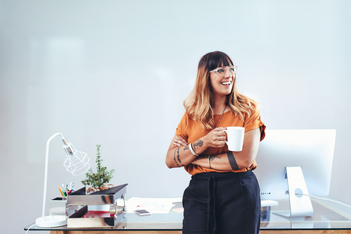Woman smiling at her desk while holding a cup of coffee. Boost your confidence through self-acceptance. 