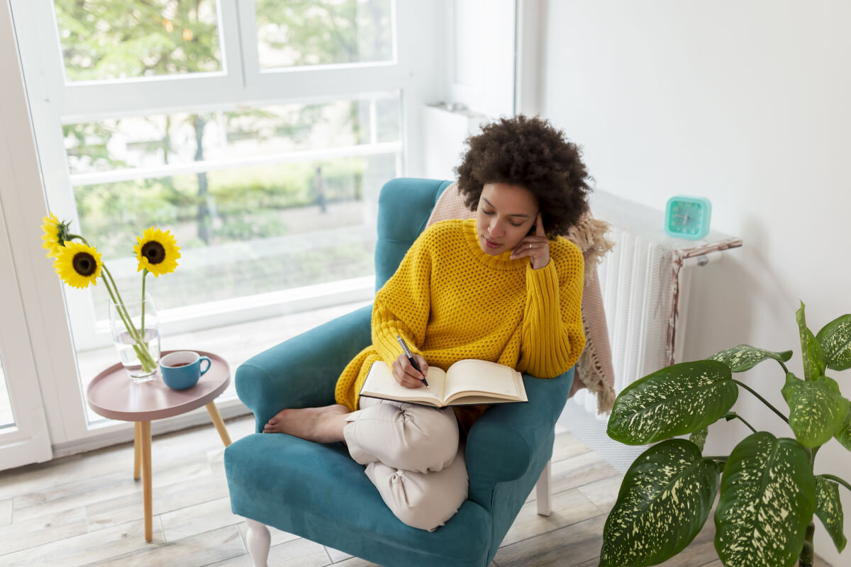 Woman in a bright yellow sweater writing in her journal with plants and sunflowers around her. 