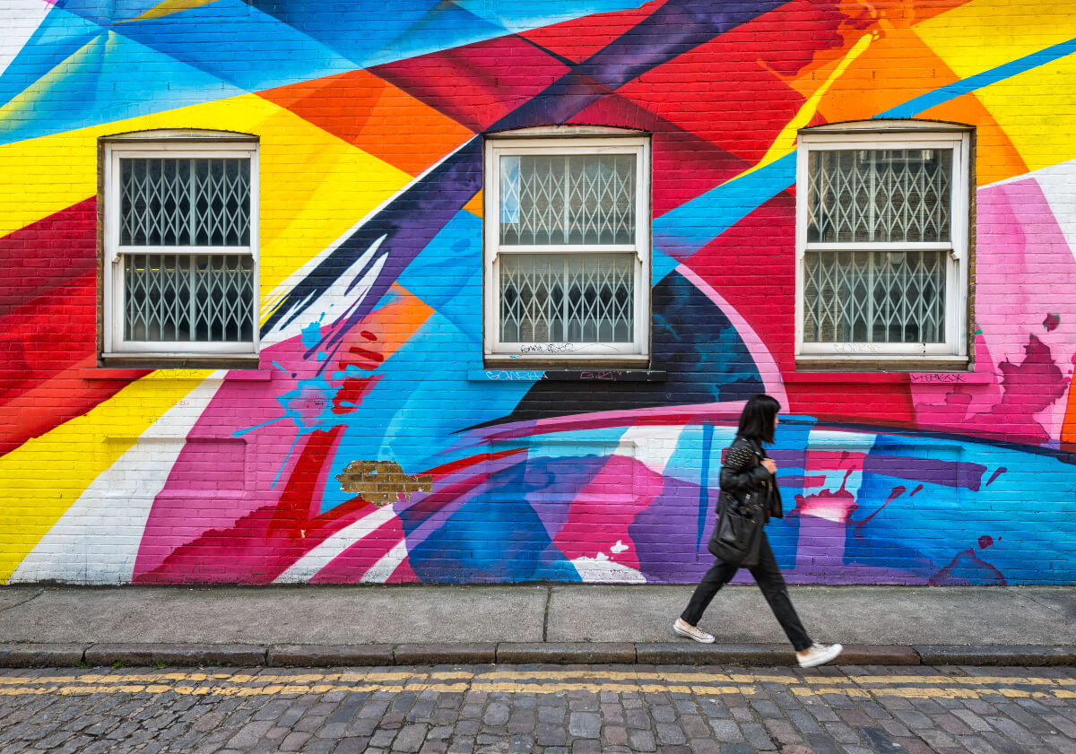 Young woman walks past a brick wall covered in colourful graffiti. Exploring the street art in your city is one way to have more art in your life.