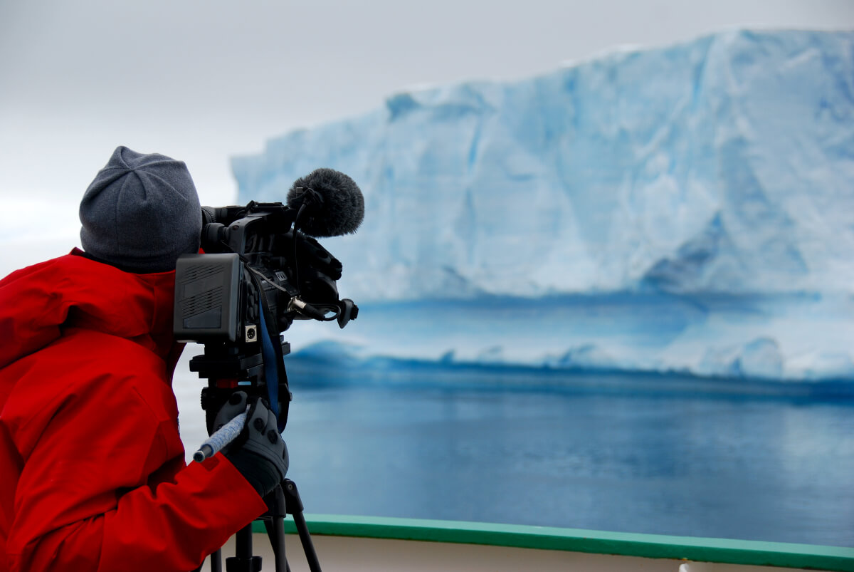 Videographer in a red coat recording video footage of an iceberg 