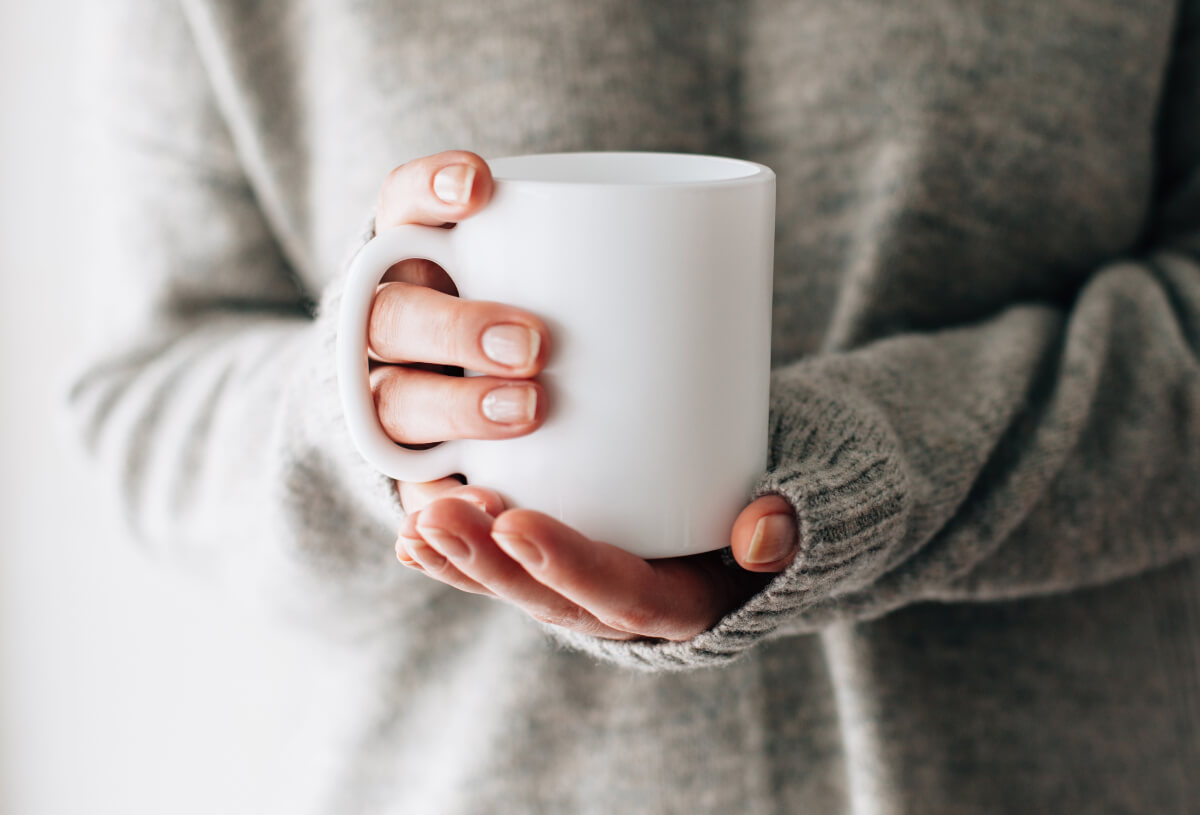 Zoomed in image of a person's hands holding a white mug. Reusable mugs and smoothie cups can be part of your sustainable morning routine. 