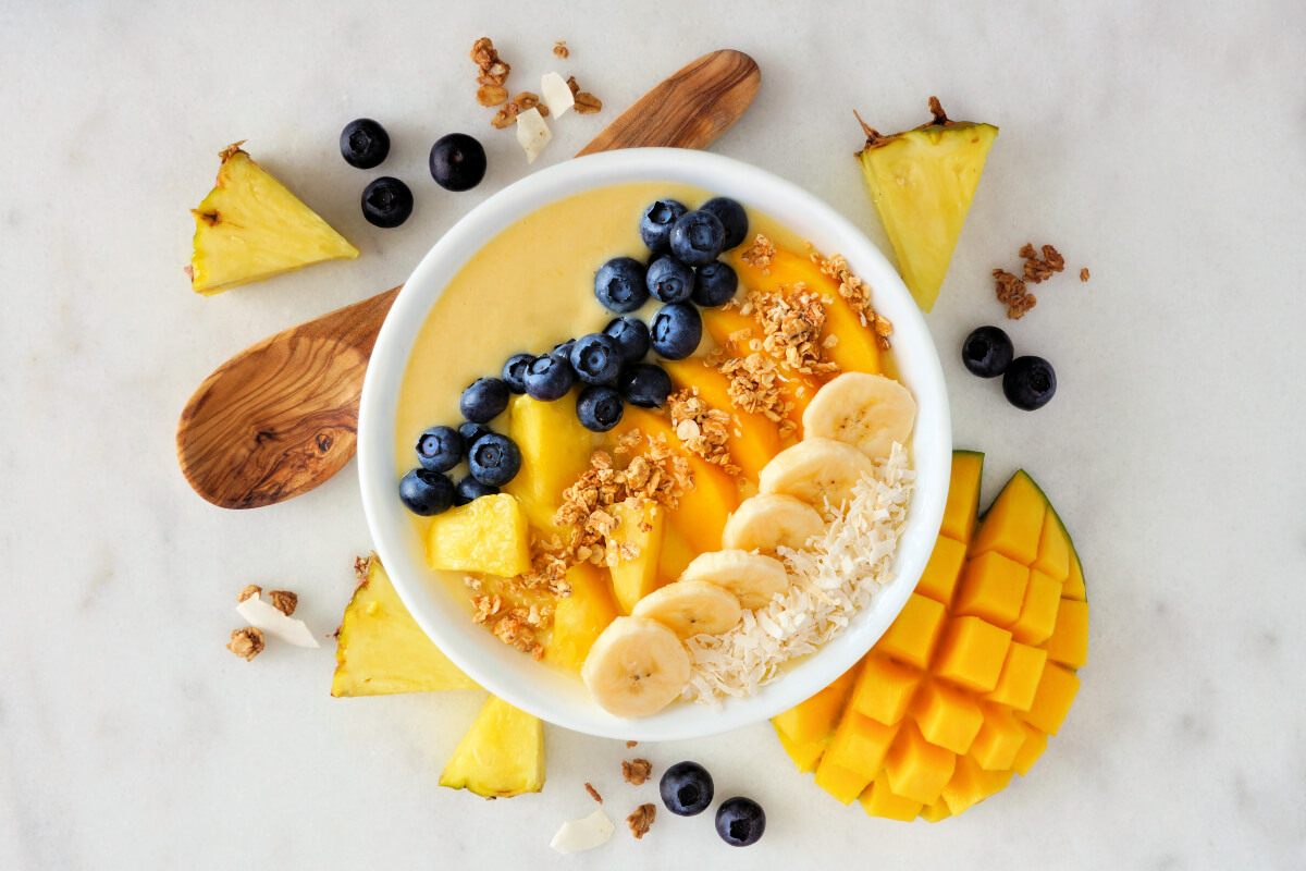 Orange smoothie bowl with mango and pineapple, topped with sliced banana, blueberries, granola, and shredded coconut. 