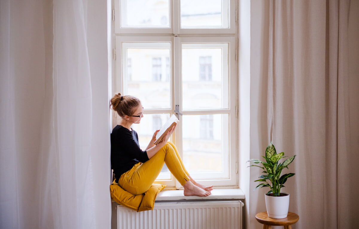 Woman in a black top and mustard yellow pants sitting on a window sill reading a book about sustainable lifestyle. 