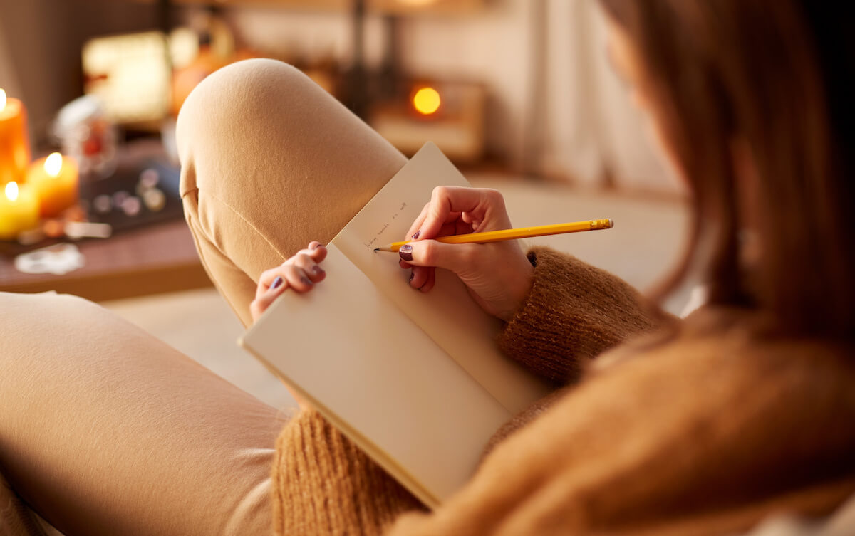 close up of young woman with pencil writing to diary and resting her feet on table at cozy home