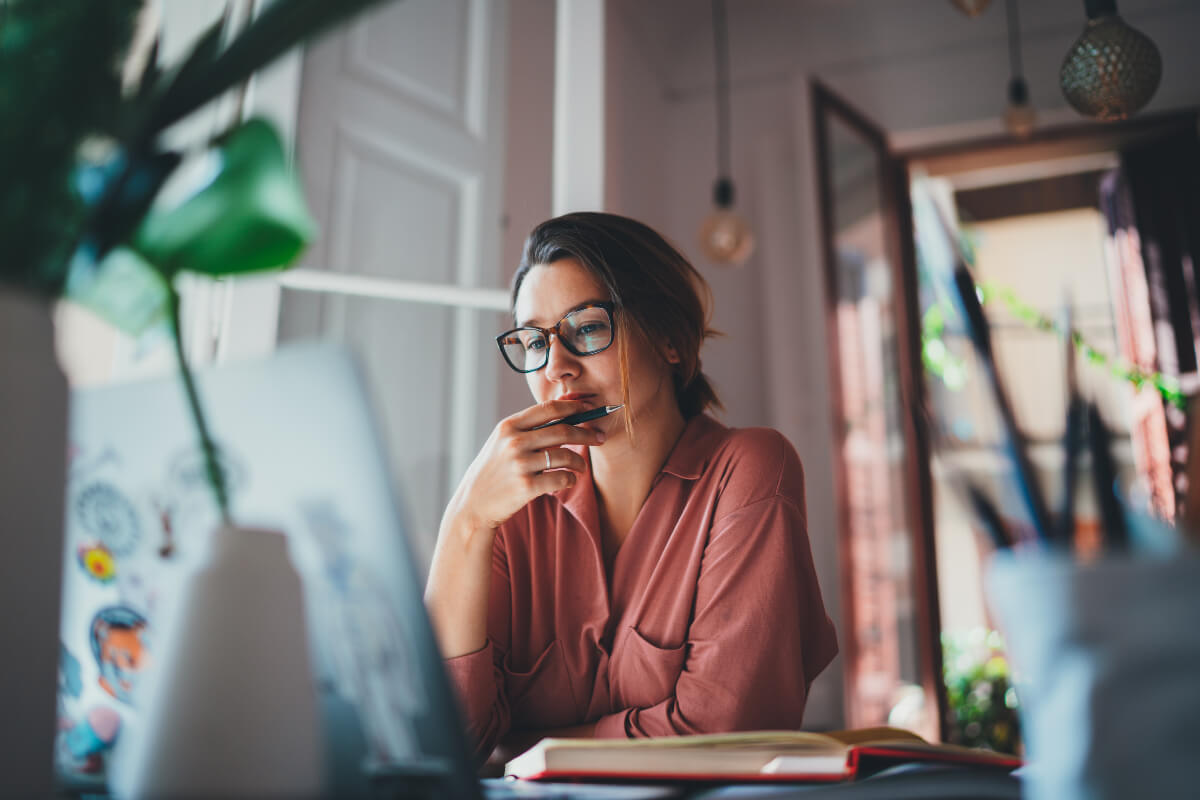 Person wearing glasses sitting at a desk surrounded by plants looking at their laptop. They are probably pondering sustainable lifestyle changes. 

