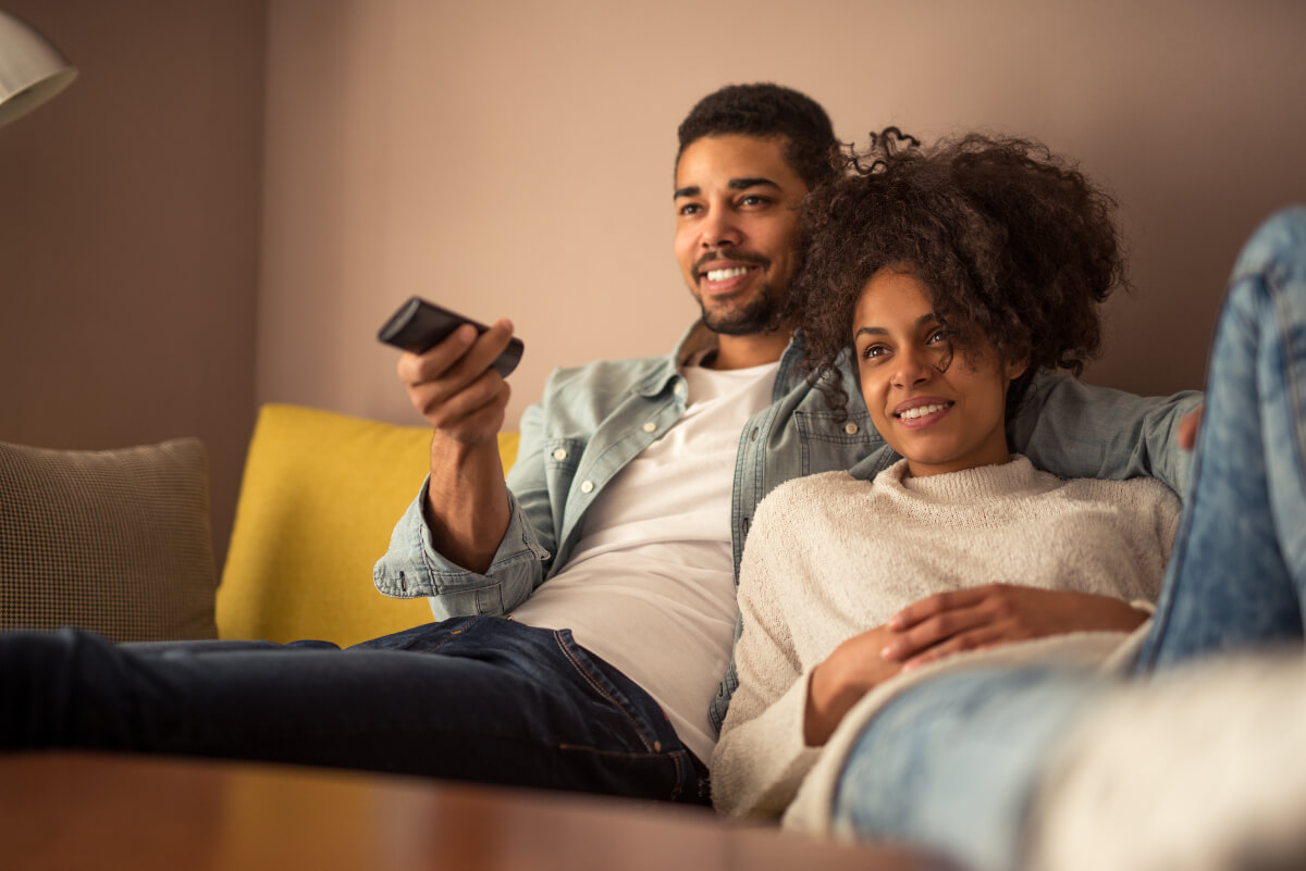 A couple sitting on a sofa watching TV with their legs comfortably outstretched. This Black History Month, check out a documentary to learn more about Black history. 