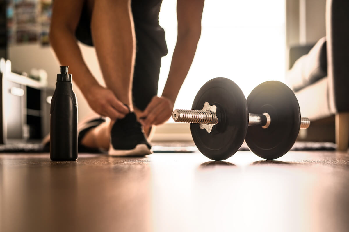 Morning workout routine in home gym. Fitness motivation and muscle training concept. Man in sneakers tying shoelaces in sunlight. Athlete starting exercise with dubbell weight.