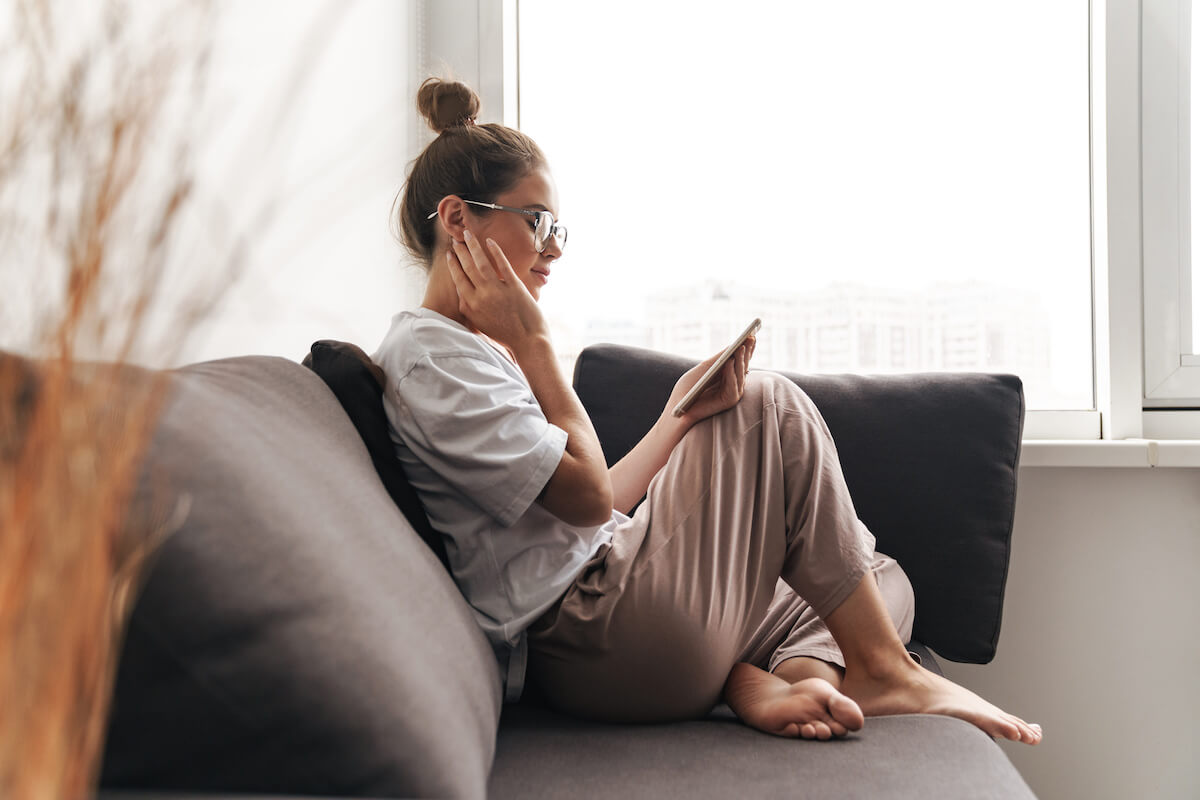 Image of concentrated nice woman in eyeglasses using cellphone while sitting on sofa at living room