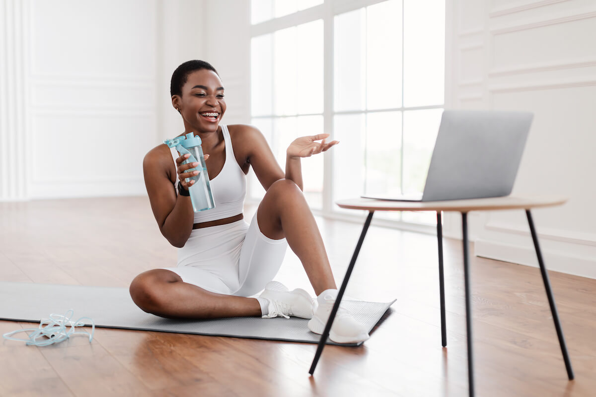 Online Exercise Session Marathon, Remote Training. Happy millennial black female sitting on mat and talking with coach, having videocall with teacher, taking break from virtual workout drinking water