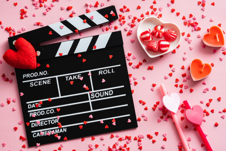 Romantic movie concept. Happy Valentines day. Clapperboard, hearts, sweets, confetti on pink table. Flat lay, top view.