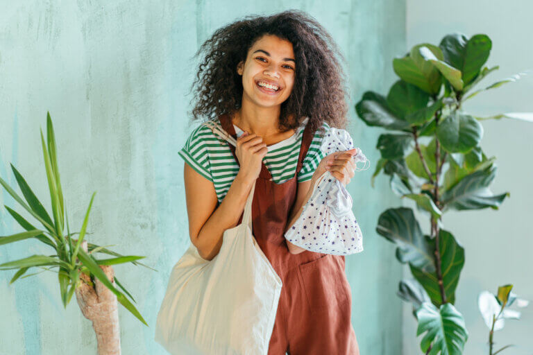 eco friendly positive mixed race woman in brown overalls and strip t-shirt wit cotton shopper with vegetables, green background.