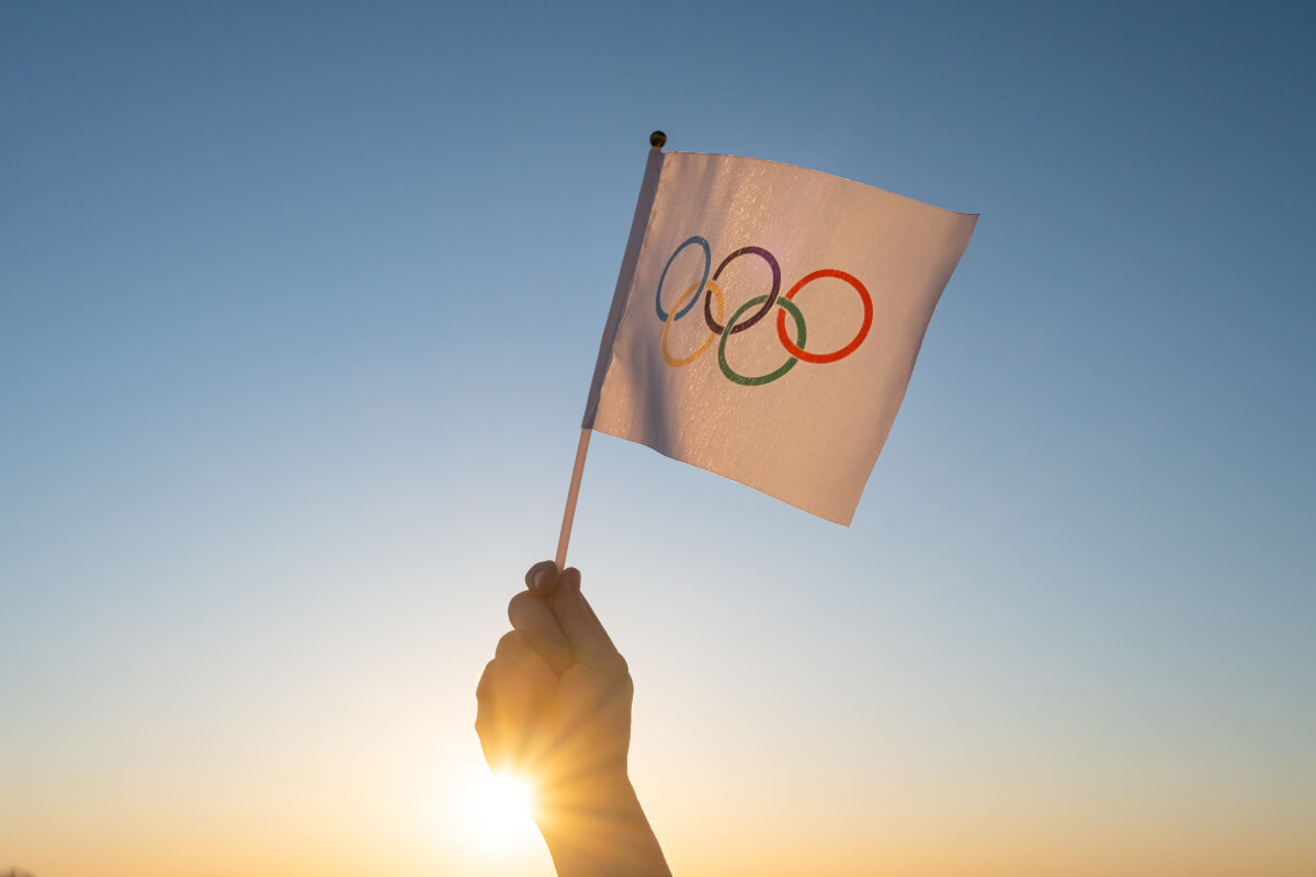 Hand holding small flag of the Olympics logo against a backdrop of the sunrise. Several countries have decided on an Olympic boycott. 