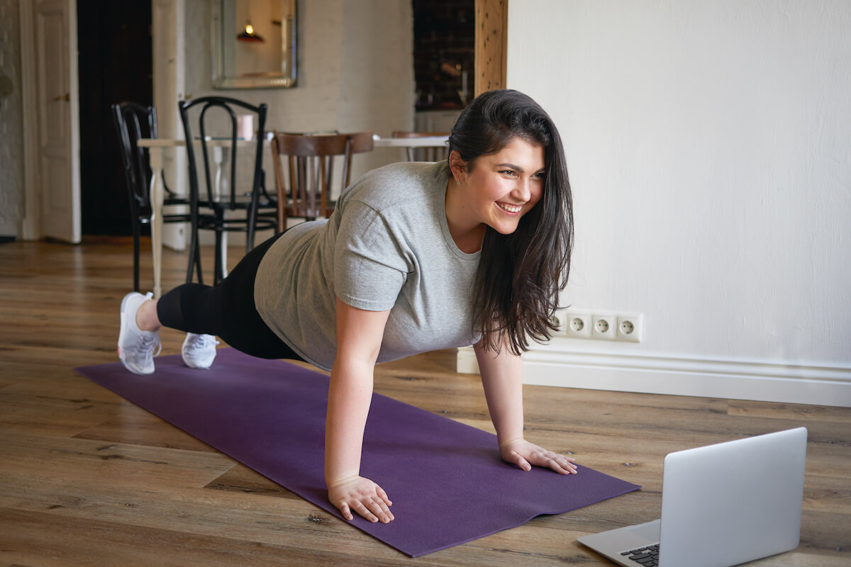 Sporty plus size woman in sportswear working out at home, doing plank on yoga mat in front of open laptop, repeating instructions by professional fitness trainer watching online video tutorial