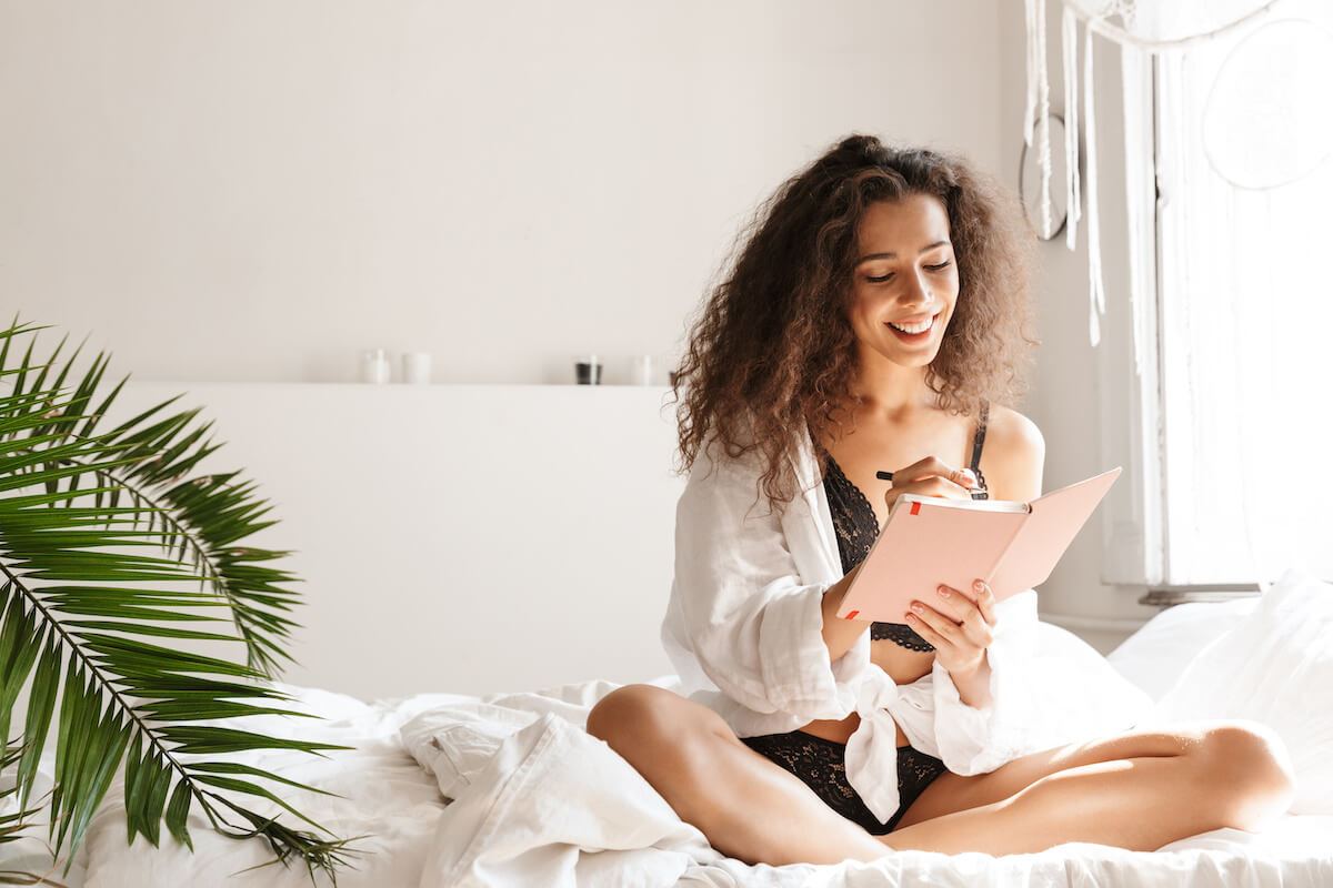 Image of adorable young woman wearing lingerie smiling and holding diary book on bed in white apartment