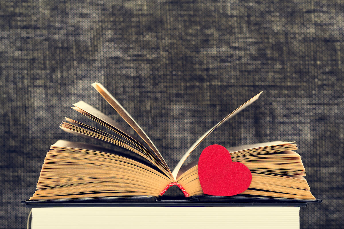 Close-up shot of open old book laying on another book, and with red heart by side, against dark colored background. Concept of love to read or reading of romance novel. Side view