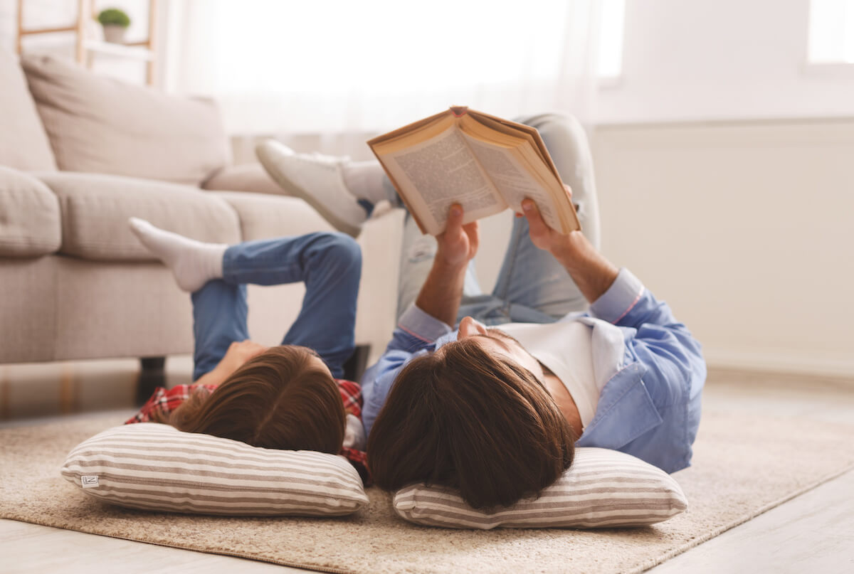 Little daughter and daddy enjoying time together, laying on floor at home, man reading book to his cute daughter, empty space