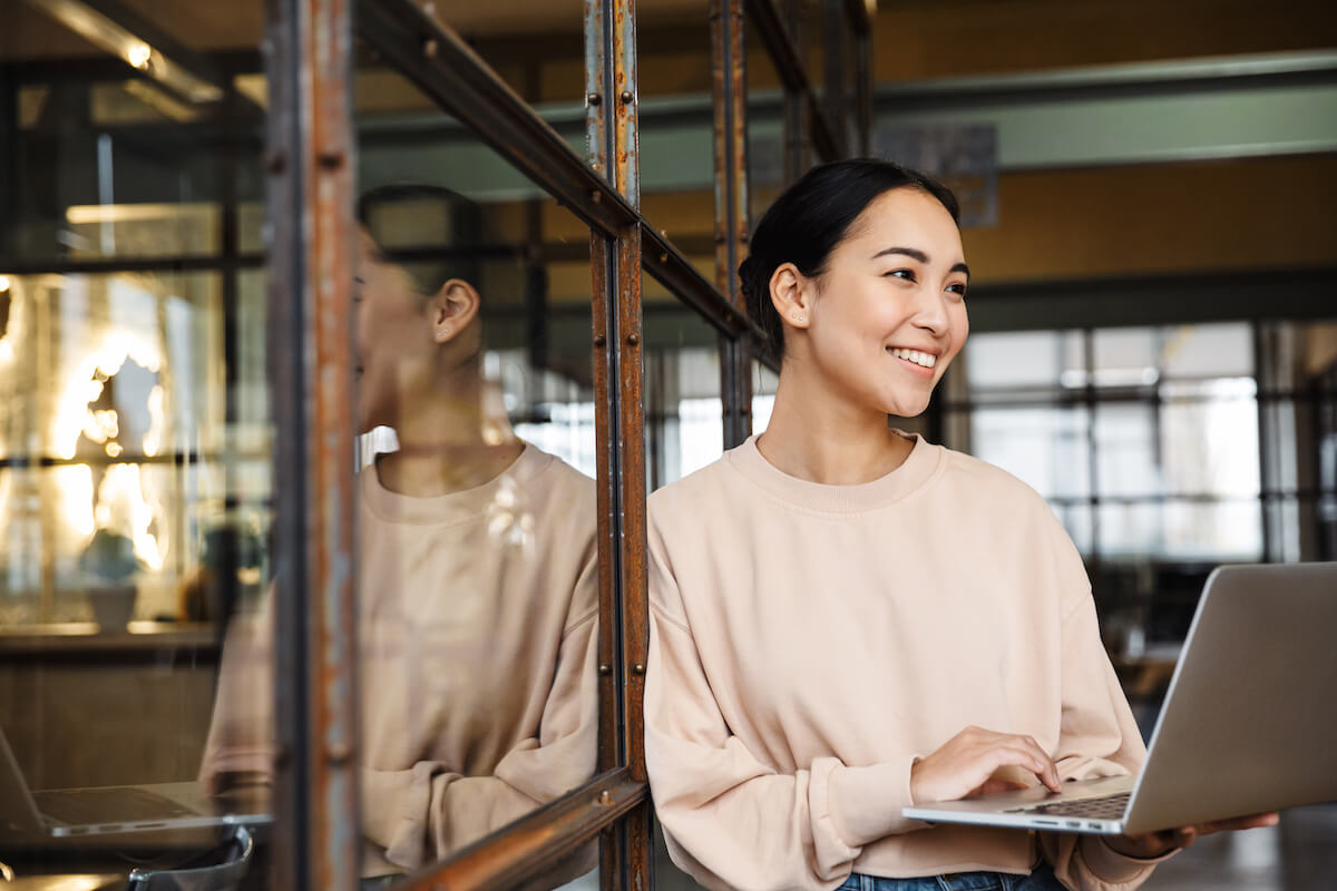 Image of young beautiful asian woman smiling and holding laptop computer while working in office