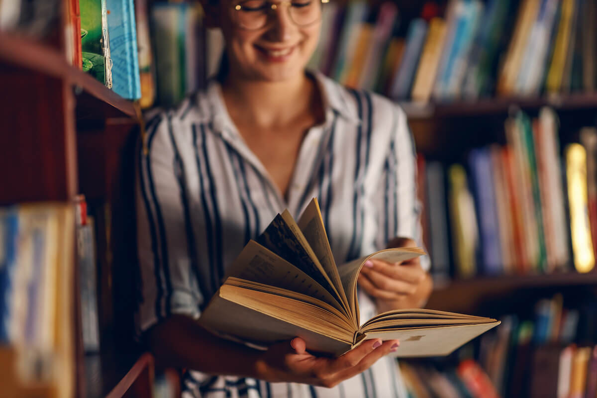 Young smiling attractive college girl leaning on book shelves in library and searching for material for homework.