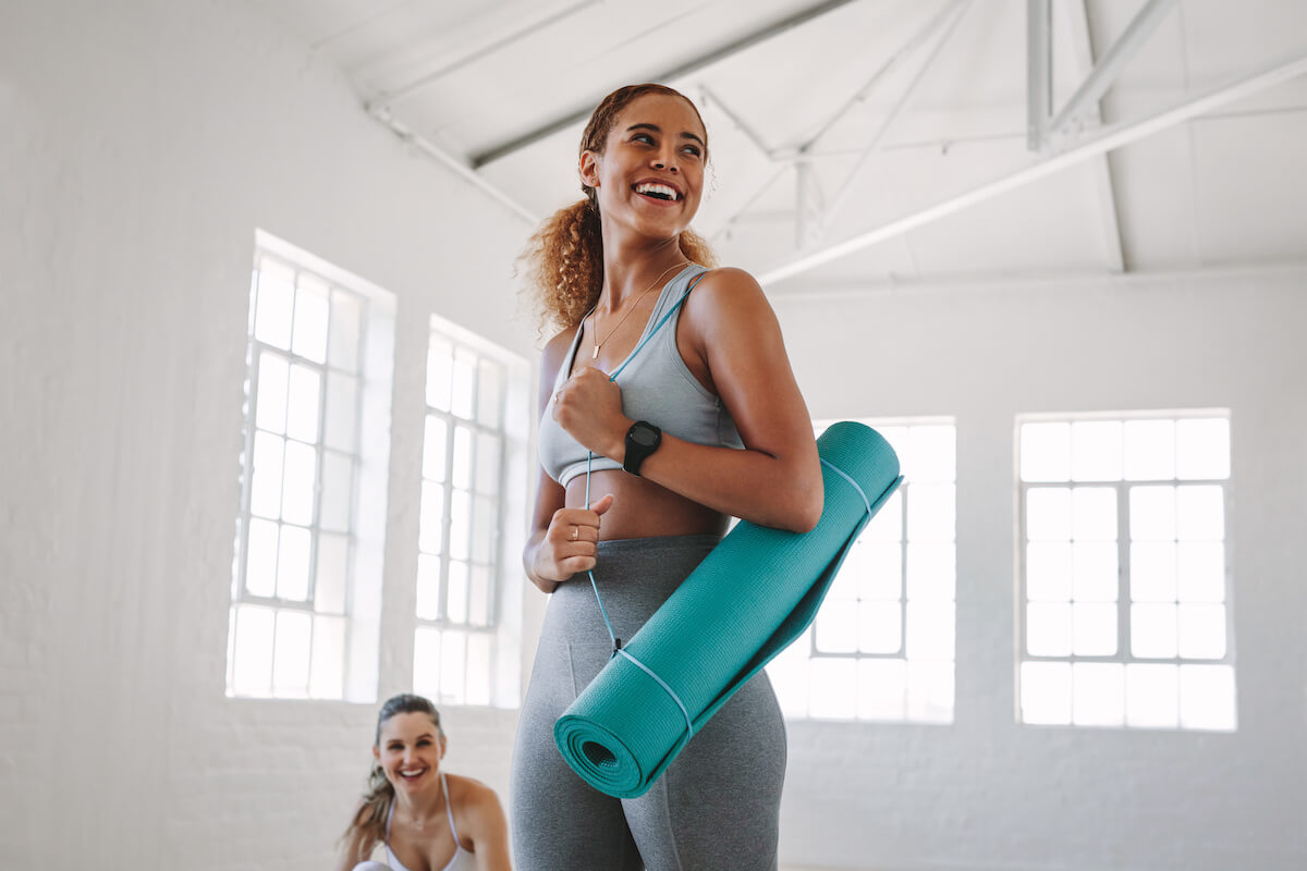 Smiling fitness woman standing in a fitness studio carrying a yoga mat. Portrait of a young woman at a fitness training centre.