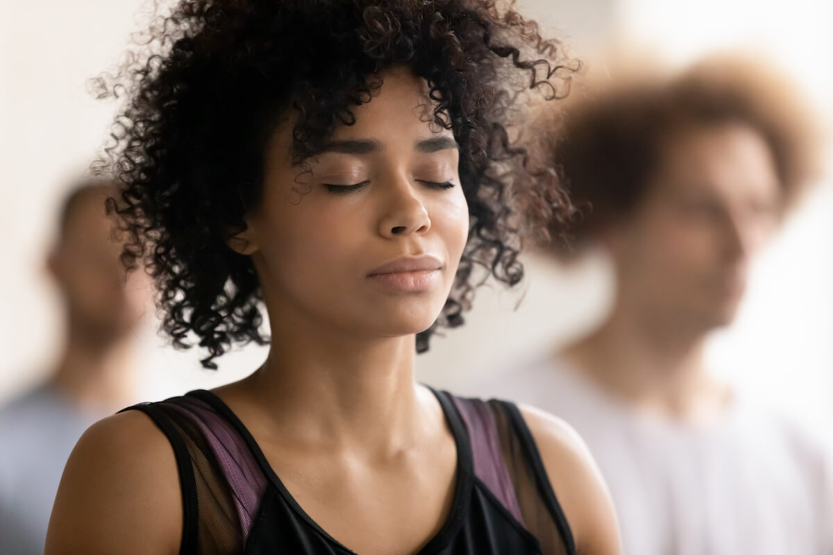 Head shot close up young peaceful attractive curly hair african american woman breathing fresh air, enjoying deep meditation with closed eyes, relaxing after yoga class workout in sport club.