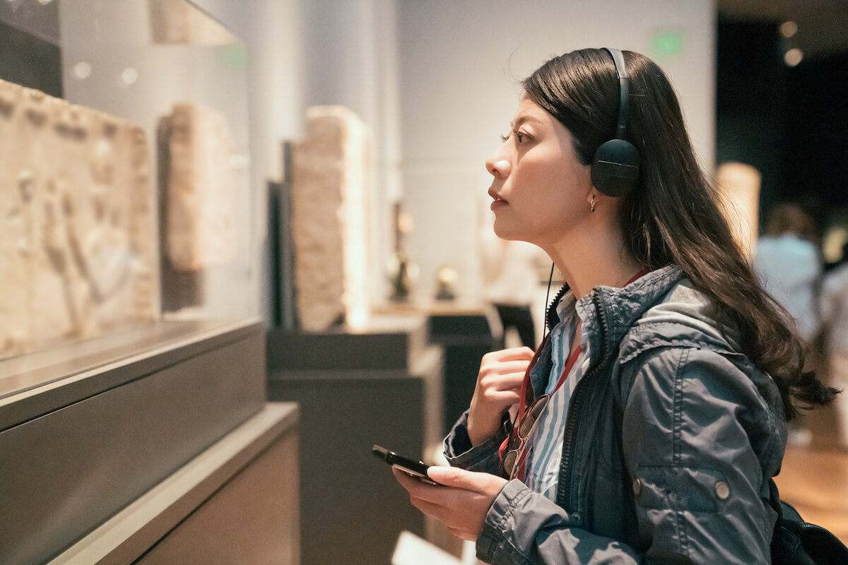 Asian woman tourist looking at exposition in museum using headphones. 
