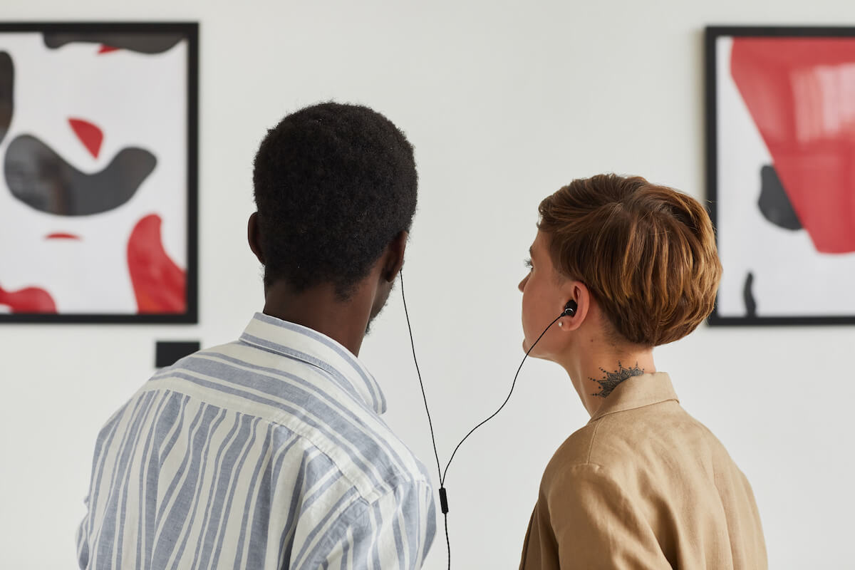Back view portrait of two young people looking at paintings and sharing audio guide while exploring modern art gallery exhibition.