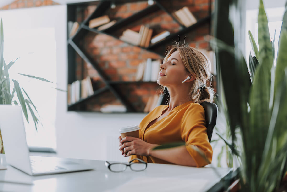 Young businesswoman sitting in the chair at her workplace and closing her eyes while holding a cup of coffee and listening to music.