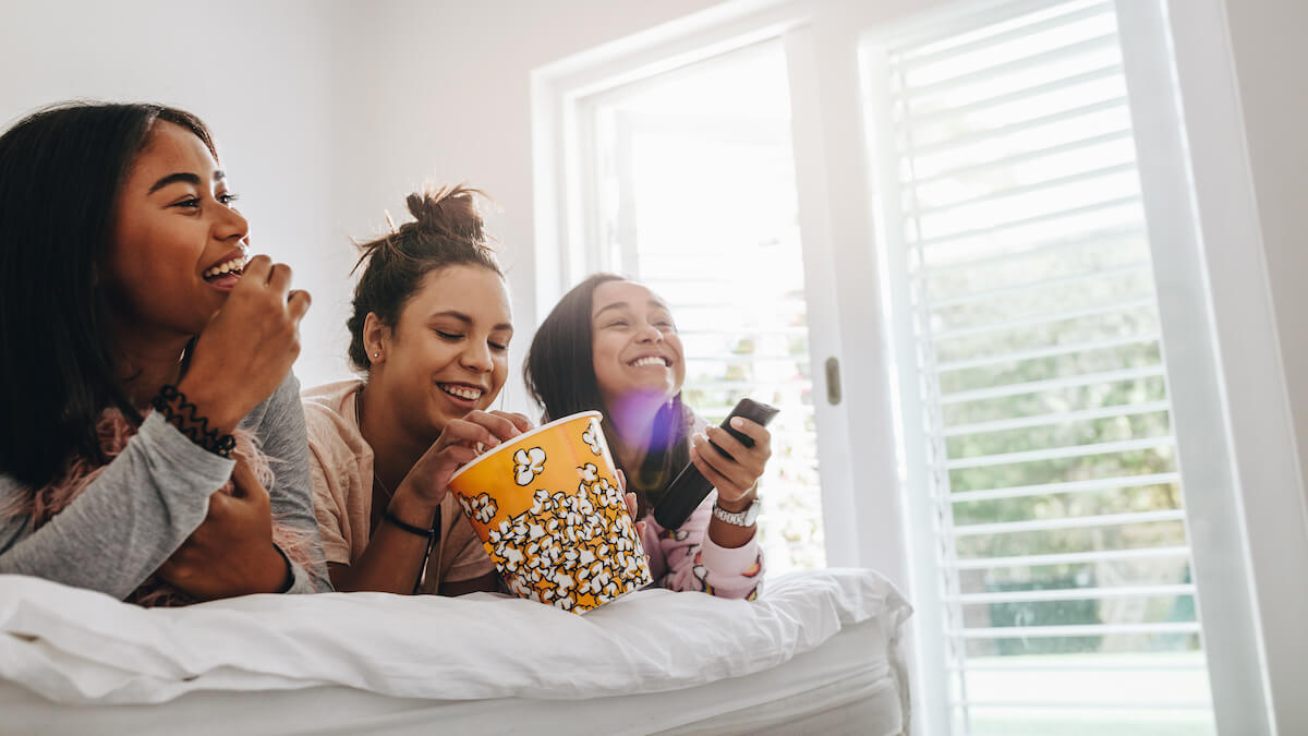 Three young girls lying on bed eating popcorns from a tub watching TV. 