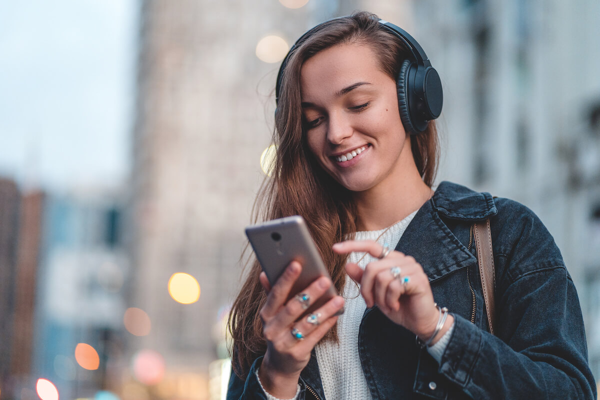 Young happy stylish trendy casual hipster woman changes songs and tracks on smartphone during listening to music on a wireless headphone while walking around the city. 