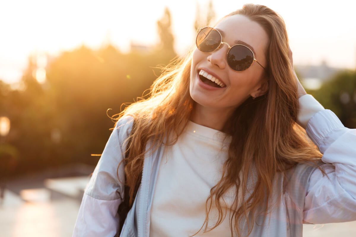 Close up picture of laughing brunette woman in sunglasses and autumn clothes posing outdoors and looking at the camera.