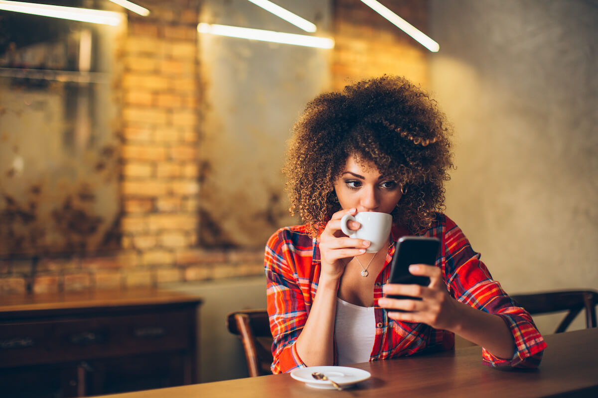 Mixed-race female looking at her cell phone while drinking coffee. 